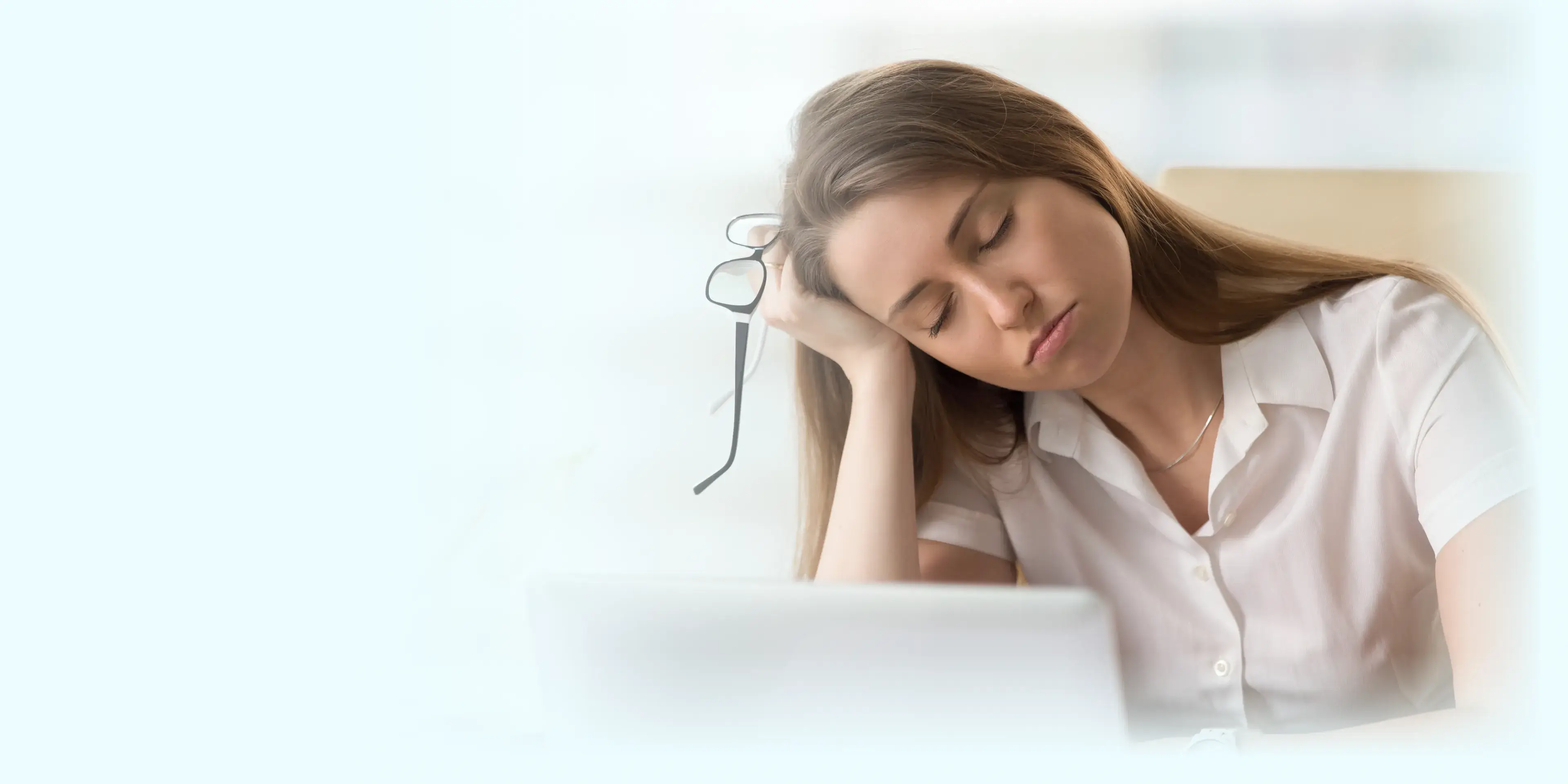 A woman is sitting at a desk, resting her head on her hand while holding glasses.